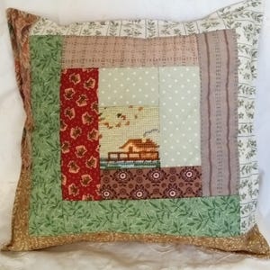 May include: A square throw pillow featuring a patchwork design with various fabric patterns including green leaves, red floral prints, and polka dots. The center panel displays a cross-stitched scene of a small house near water.