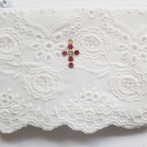 May include: White lace pouch with a zipper closure. The pouch is adorned with a cross made of red and gold rhinestones. The lace features a floral pattern.