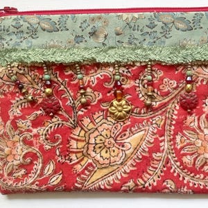 May include: A rectangular, red paisley-patterned pouch with a green floral top and a red zipper. The pouch is adorned with green fringe and beaded embellishments. A small fabric loop is attached to the side.