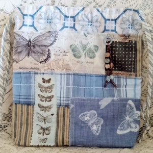 May include: A patchwork tote bag with a variety of blue and white fabrics. The bag features butterfly and floral patterns, with a drawstring closure and a beaded accent. The bag's design includes a mix of stripes, plaid, and printed butterfly motifs.