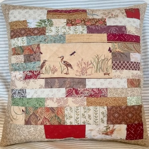 May include: A square patchwork throw pillow featuring a central panel with illustrations of two herons, a dragonfly, and a turtle amidst reeds. The surrounding border consists of various rectangular fabric strips in shades of red, tan, green, and brown with floral, paisley, and geometric patterns.