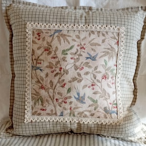 May include: A square throw pillow featuring a central panel with a botanical print of blue birds, green leaves, and red berries on a cream background, framed by cream-colored lace trim and a beige and white checkered border fabric.