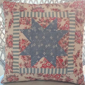 May include: A square throw pillow featuring a central blue fabric star with a white floral pattern. The star is framed by a light blue and white striped border, set against a cream-colored background with a red and gray floral print pattern.