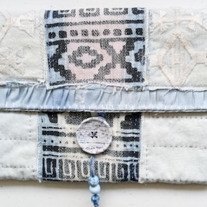Puede incluir: Un bolso de mano hecho a mano con un diseño de patchwork. La solapa superior presenta un volante azul claro y un cierre de botón. El bolso está hecho de varias telas, incluyendo mezclilla con patrones geométricos y tela blanca acolchada.