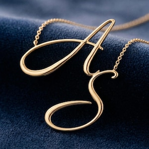 May include: A gold-coloured necklace featuring the cursive letters "A" and "S". The pendant hangs from a delicate chain, set against a dark blue background. The letters are in a flowing script style.