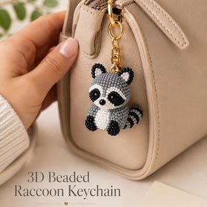 May include: A 3D beaded raccoon keychain featuring a grey, white, and black color palette. The raccoon charm is attached to a gold-toned metal chain and clasp. The text 3D Beaded Raccoon Keychain Finished Charm is visible in the image.