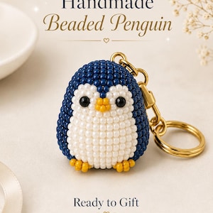 Può includere: Un portachiavi a forma di pinguino realizzato a mano con perline blu scuro, bianche e gialle e occhi neri. Il testo "Handmade Beaded Penguin" e "Ready to Gift" è visibile sopra e sotto il pinguino, fissato a un anello in metallo dorato.