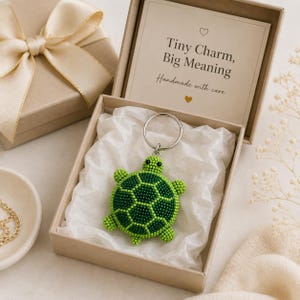 May include: A beaded turtle keychain in shades of green with black eyes, resting on white tissue paper inside a square cardboard gift box. The box lid features the text Tiny Charm, Big Meaning and Handmade with care, decorated with a cream ribbon.