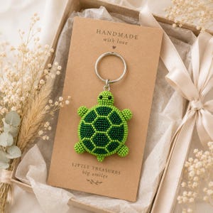 May include: A beaded turtle keychain featuring a green and dark green shell pattern, attached to a silver metal ring. The keychain is mounted on a rectangular tan card with the text "HANDMADE with love" and "LITTLE TREASURES big smiles".