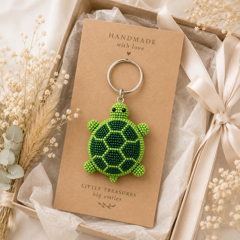 May include: A beaded turtle keychain featuring a green and dark green shell pattern, attached to a silver metal ring. The keychain is mounted on a rectangular tan card with the text "HANDMADE with love" and "LITTLE TREASURES big smiles".