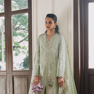 Op de afbeelding: Een lichtgroene geborduurde ensemble met een tuniek met lange mouwen, bijpassende broek en een doorschijnende sjaal. De outfit is versierd met bloemen en geometrische patronen in roze en wit. Een kleine paarse tas en gouden sieraden maken de look compleet.