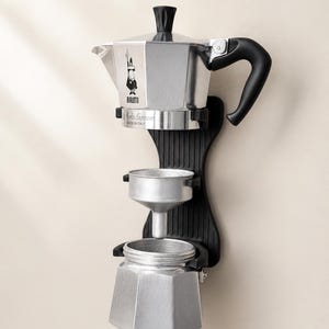Pode incluir: Uma cafeteira italiana prateada com uma alça preta e uma base preta. A cafeteira tem um design clássico com um pequeno logotipo e as palavras "Moka Express" impressas na lateral. A cafeteira é fabricada na Itália.