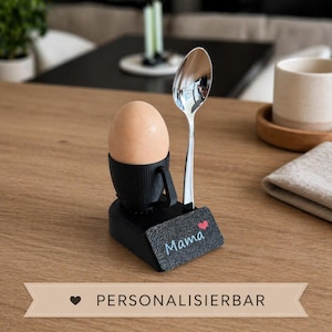 Könnte beinhalten: Ein schwarzer Eierbecher mit Henkel, der ein braunes Ei und einen silbernen Löffel hält. Auf der Basis steht das Wort "Mama" mit einem Herz. Der Text "PERSONALISIERBAR" steht unten.