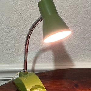 May include: A desk lamp with a glossy olive green base and a matching cylindrical shade. The lamp features a flexible silver-toned metal gooseneck arm and a white power switch located on the front of the base. The light is illuminated.