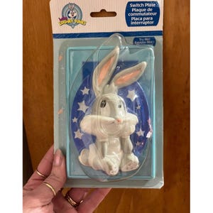 May include: A Bugs Bunny switch plate in a plastic package. The gray and white Bugs Bunny figure is centered on a blue background with white stars. The package has the Looney Tunes logo and text in English, French, and Spanish.