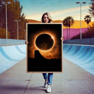 Può includere: Una stampa artistica da parete rettangolare incorniciata raffigurante un'eclissi solare con la sagoma di una persona in bicicletta che salta su una rampa. L'opera mostra un sole circolare scuro circondato da una corona arancione brillante e il testo CHASE THE LIGHT.