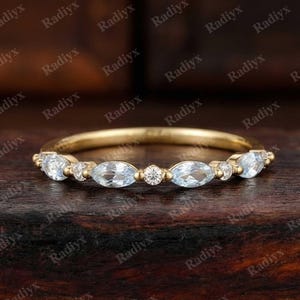 May include: A gold band ring featuring alternating light blue marquise-cut gemstones and small round white gemstones. The ring is set on a dark wooden surface, showcasing the ring's design and sparkle.