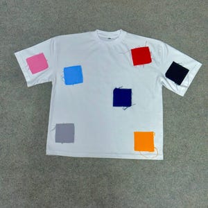Puede incluir: Camiseta blanca con seis parches cuadrados de colores cosidos. Los parches son rosa, azul, rojo, negro, gris y naranja. Los parches están cosidos con hilo visible.