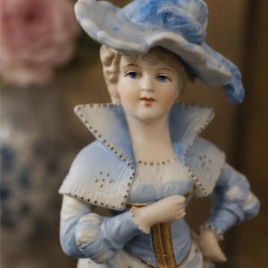May include: A porcelain figurine of a person wearing a wide-brimmed blue and white hat, a high-collared blue jacket with gold trim, and a white skirt. The figure has painted facial features, blue eyes, and a hand resting on the waist.