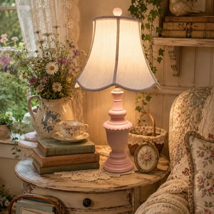 May include: A table lamp with a light pink pedestal base and a flared, off-white fabric lampshade. The lamp features a smooth, tiered column design with a small spherical finial at the top, emitting a warm, soft glow from within the shade.