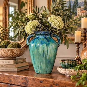 May include: A ceramic vase with a marbled blue and green glaze featuring two side handles. The vase is filled with white hydrangea flowers and green foliage, positioned on a wooden surface next to books and a speckled bowl.