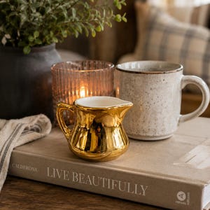 May include: A small, shiny gold-toned ceramic creamer pitcher with a curved handle and spout, positioned next to a speckled beige ceramic mug and a ribbed glass candle holder on top of a book titled Live Beautifully by Calderone.