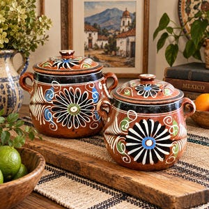 May include: Two round terracotta pots with lids, each featuring two side handles and hand-painted floral patterns in black, white, blue, and green. The pots have a dark brown band around the upper rim and a glossy finish on the exterior surface.