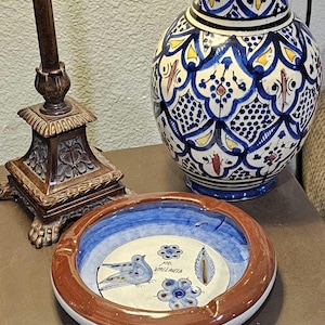 May include: A decorative ceramic ashtray and vase. The ashtray has a brown rim, blue and white interior with a bird and floral design. The vase is blue and white with a detailed pattern. The ashtray has the text "Puerto Vallarta".