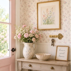May include: A light-colored wooden console table holds a ceramic pitcher filled with pink flowers, a scalloped bowl, and a framed sign that reads home sweet home. A gold key-shaped wall hook with hanging keys is mounted on floral wallpaper.