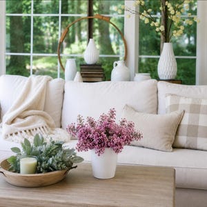 May include: A white ceramic vase filled with purple lilac flowers sits on a wooden coffee table next to a wooden bowl containing a green plant and a candle. A white sofa with a cream throw blanket and a beige plaid pillow is in the background.