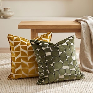 May include: Two decorative cushions. One is mustard yellow with a repeating geometric design. The other is olive green with a stone-like pattern. Both cushions are on a cream-coloured rug.