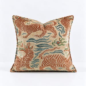 Puede incluir: Un cojín cuadrado con base de terciopelo beige y un patrón repetitivo de tigres estilizados en naranja quemado y azul apagado. El diseño incluye motivos en forma de nubes arremolinadas y bordes decorativos con textura.
