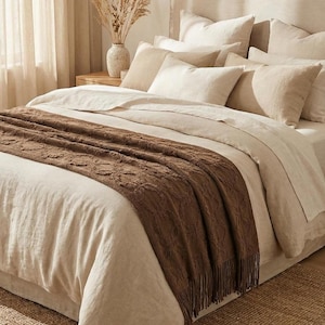 May include: A bed with beige linens and pillows, topped with a brown patterned throw blanket with fringe. The bed is in a well-lit room with a neutral color palette, creating a cozy and inviting atmosphere. A vase with dried plants sits on a wooden nightstand.