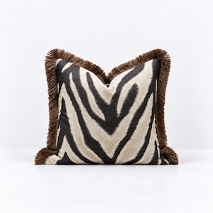 May include: A square throw cushion featuring a black and beige zebra stripe pattern. The cushion is finished with a thick, dark brown fringe trim along all four edges, creating a textured border around the central animal print fabric design.