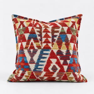 May include: A square throw pillow featuring a woven geometric pattern in vibrant shades of red, blue, yellow, and green against an off-white background. The design consists of repeating triangular and diamond shapes across the entire surface.