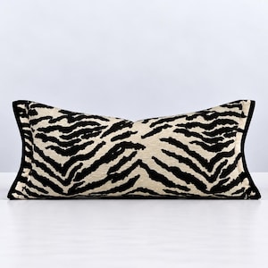 Puede incluir: Un cojín lumbar rectangular con un patrón de rayas de cebra en negro y crema. El cojín tiene una forma curva distintiva en los lados izquierdo y derecho, acabado con un ribete negro sólido a lo largo de los bordes de la funda de tela.