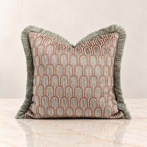 May include: A square throw pillow featuring a repeating arch pattern in muted pink and blue-grey tones. The pillow is finished with a thick, textured sage green fringe trim along all four edges, creating a decorative border around the fabric.