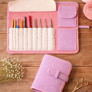 Puede incluir: Un estuche de ganchillo hecho a mano en rosa, lavanda y blanco. El estuche abierto muestra varios ganchillos en un bolsillo blanco. Un libro de ganchillo lavanda a juego está cerrado con una correa. Unas tijeras doradas descansan sobre la superficie de madera.