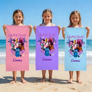 Op de afbeelding: Drie strandlakens in roze, paars en blauw worden door drie jonge meisjes omhoog gehouden. Elk laken heeft een afbeelding van een groep vrouwen, een blauwe kat en de naam "Emma" eronder. Het woord "HUNTRAX" staat bovenaan gedrukt.