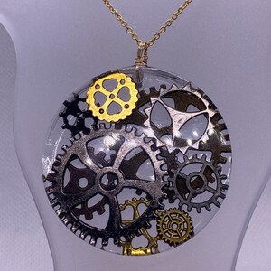 May include: A gold chain necklace with a round pendant featuring a cluster of silver and gold gears encased in clear resin.