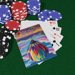 Puede incluir: Una baraja de cartas con un diseño de globo aerostático colorido, junto a pilas de fichas de póquer rojas, azules y negras sobre una superficie verde. Cuatro cartas están abanicadas, mostrando el diez de corazones, diamantes, picas y tréboles.