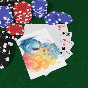 May include: A deck of playing cards with a whimsical illustration of a blue creature expelling colorful bubbles. Poker chips in red, black, and blue are scattered around the cards on a green surface. The cards show the number 10.
