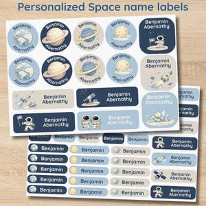 Op de afbeelding: Vellen gepersonaliseerde naamlabels met ruimtethema. Ontwerpen omvatten planeten, astronauten, raketten en de tekst "Benjamin Abernathy". De labels zijn in tinten blauw, beige en wit.