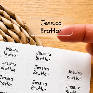 May include: A clear rectangular adhesive label with the name Jessica Brattan printed in black sans-serif font. The label is held against a sheet of identical white rectangular labels, each featuring the same name in a black sans-serif typeface.