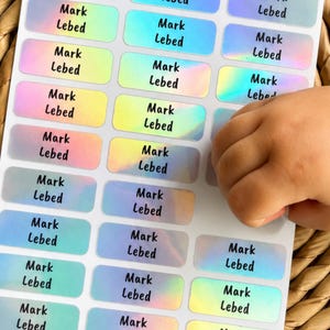 Puede incluir: Una hoja de etiquetas adhesivas rectangulares con un acabado holográfico iridiscente. Cada etiqueta presenta el nombre Mark Lebed impreso en una fuente negra, negrita y sin remates. Las etiquetas están dispuestas en una cuadrícula sobre papel blanco.
