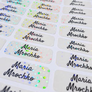 Op de afbeelding: Een vel met rechthoekige zelfklevende labels waarop de naam Maria Mrochko in een zwart cursief lettertype is gedrukt. Elk label heeft een semi-transparante afwerking met een verspreid holografisch glinstereffect dat licht reflecteert.