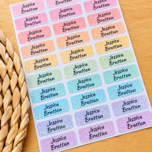 May include: A sheet of rectangular adhesive labels featuring a gradient of colours from pink to purple. Each label has a holographic dot pattern and the name Jessica Brattan printed in a black cursive font. The labels are arranged in a grid.