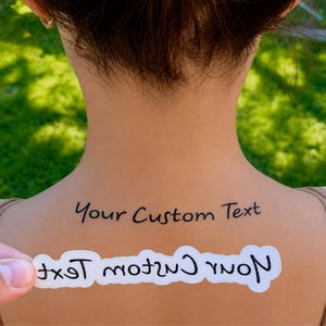 May include: A temporary tattoo applied to the skin featuring the black text Your Custom Text in a handwritten font style. Below the applied tattoo, a white paper backing strip displays the same text in a mirrored, reversed orientation.