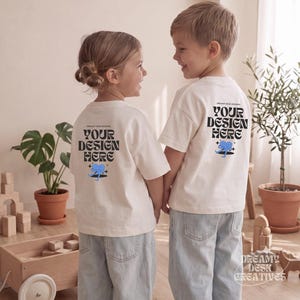 May include: Two off-white short-sleeve t-shirts featuring black text that reads YOUR DESIGN HERE in a wavy font, accompanied by a small blue graphic of a character and black stars. The shirts are worn by two children facing each other.