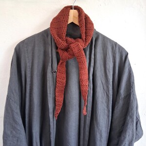 May include: A dark charcoal grey open-front jacket with long sleeves and a relaxed fit, layered with a rust-coloured crocheted scarf tied in a simple knot. The scarf features a textured, repeating stitch pattern and tapered ends, draped over a wooden hanger.